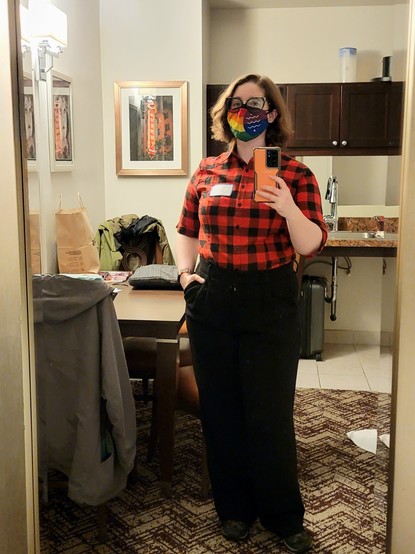 A selfie of Erin, a white non-binary person with shoulder length dark blonde hair. They are wearing a red and black plaid shirt tucked into high waisted black slacks and a Bellingham pride flag mask.