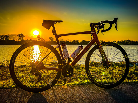 A photo of a red BMC Roadmachine road bike standing next to the Welland Canal at sunrise. The sun is rising over the rear wheel of the bike and it’s reflection can be seen in the water through the spokes of the rear wheel. 