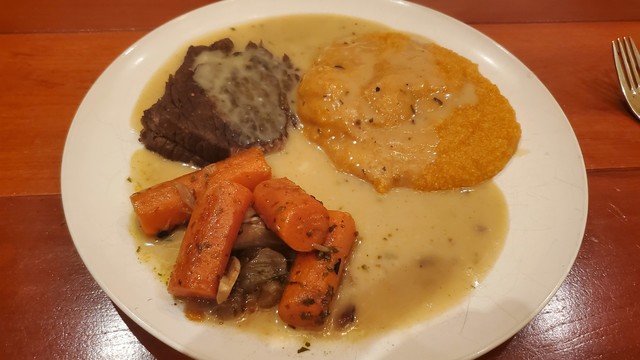 A white plate on a reddish wood table. The plate has a slice of roast beef in the upper left, some reddish polenta in the upper right, and carrots in the lower left - all covered in gravy. 