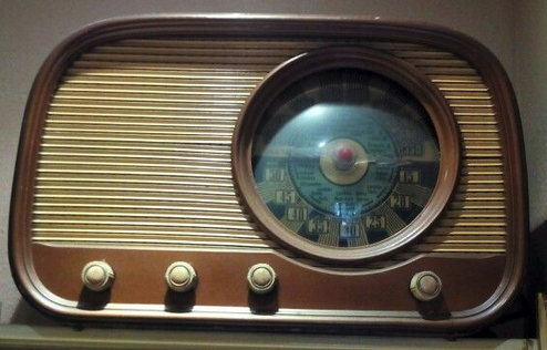 A two tone vintage radio has a strong round dial on the right, and a pale gold grille, with four knobs along the bottom