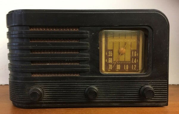 A deep-brown vintage radio, probably an early one from the 20s, has a rounded-corners square dial on the right, and a series of five horizontal lines for a grille along the left.