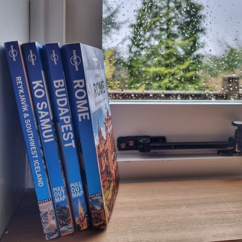 Four Lonely Planet pocket guidebooks (Reykjavik,  Ko Samui, Budapest and Rome) on a window sill. There are raindrops on the window pane and a tree in the distance.