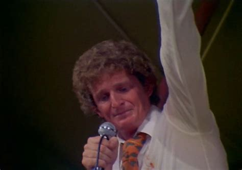 A young curly-haired man in a white shirt and wide orange tie, holding a microphone in one hand, his other arm raised in the air