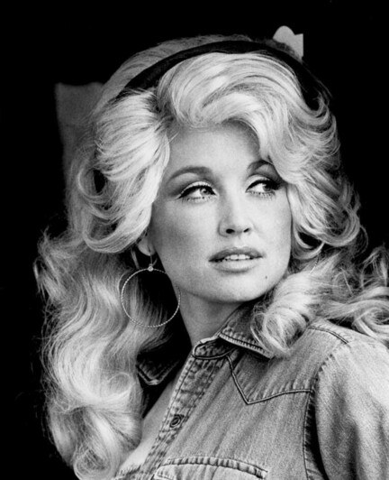 A black and white photo of Dolly Parton, taken when she was in her twenties. She has long, wavy blonde hair, and is wearing a denim shirt and large hoop earrings. Dolly is looking at something off to the side.