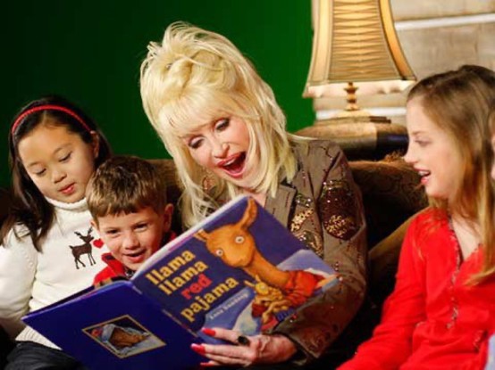 A promotional color photo of Dolly Parton reading the book "llama llama red pajama" to three children. She has blond hair, a tan jacket, and her mouth is open as she reads from the book. Three children around her look at the book as she reads.