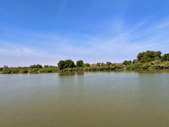 River Nile in Khartoum - the water is greenish. The skies a brilliant blue. There are trees along the river bank. 