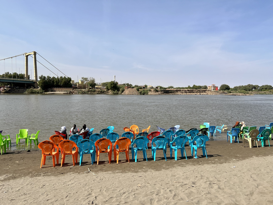 Colourful plastic chairs on the sand bank of the river Nile. Three people have moved their chairs in the shallow water. Great place to drink coffee. 