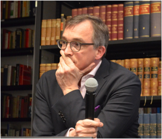 Ein Foto von Andreas Rödder in der Bibliothek des Konservatismus. Er steht mit einem Mikrofon in der einen Hand vor einem Bücherregel. Die andere Hand hat er über seinen Mund gelegt und seine Augen sind weit aufgerissen.
