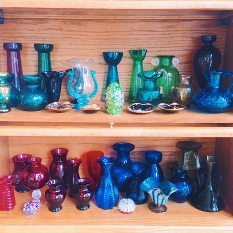 Two shelves filled with glass hyacinth vases. On the top shelf the vases are in shades of blue and green, and the bottom shelf has vases of red and blue. A couple of glass paperweights are tucked in among the vases. The vases are used primarily for hyacinth forcing in the winter months.