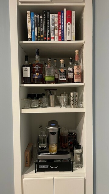 A home bar/bookshelf with storage at the top and bottom; the Bartesian and capsule drawer on the lowest shelf, glassware above it, bottles of whiskey above that, and cookbooks on the top shelf.