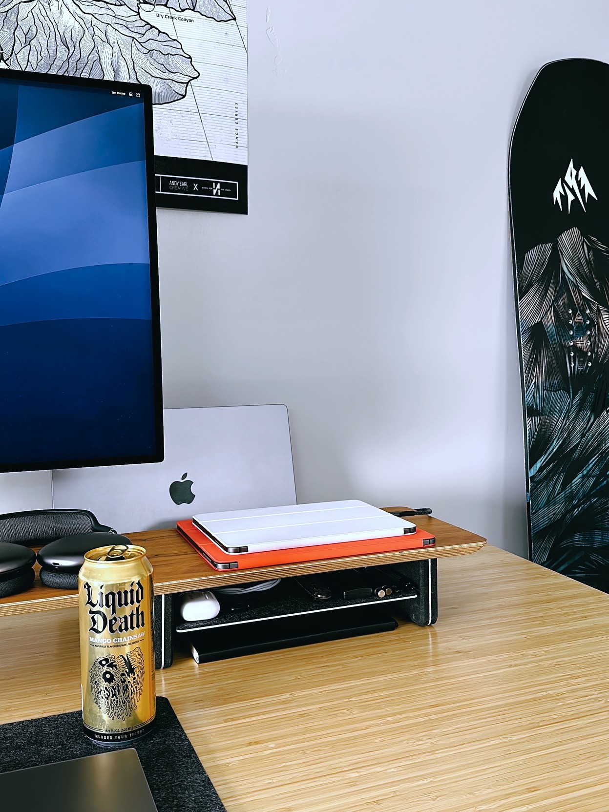 Photograph of a work from home desk setup. Pictured: part of a Pro Display XDR, AirPods Max, MacBook Pro, two iPad Pros, a Liquid Death Mango Chainsaw, and a Jones brand snowboard neatly arranged on a desk.