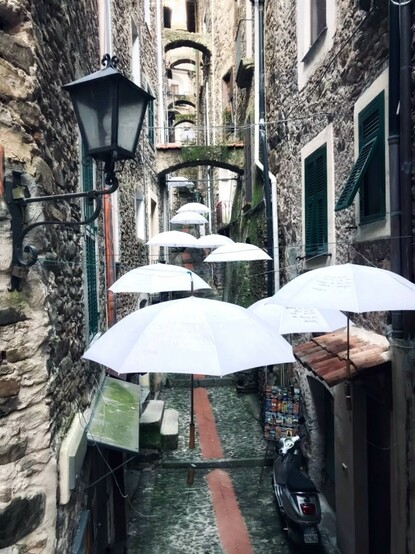 Portrait size picture showing a narrow dark alley wide enough for barely two people, between tall several story buildings. Several white open umbrellas hang overhead. 