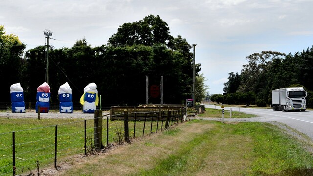 A photo of the entrance to a dairy, taken from State Highway 1 (the main road from Cape Reinga to Bluff, running the length of New Zealand).

Alongside the driveway are eight baleage rolls - a bit like giant lego heads, stacked in twos.  Each is blue and has a hat on it.  Oh and a face, so they look like Smurfs.  The four Smurfs from left to right are one that looks like Sleepy Smurf, Papa Smurf, Brainy Smurf and Smurfette.  

To the right can be seen the entrance to the farm from State Highway 1; barrelling southward and on the right side of the road from this vantage point, is a white truck and double trailer .  It has two tall covered bins - maybe carrying something like stockfeed.  Or chocolate chips.

Being a farm, there are also several fences and other roadside infrastructure, including a power pole and sign.

Oh and there is grass.  It is green.