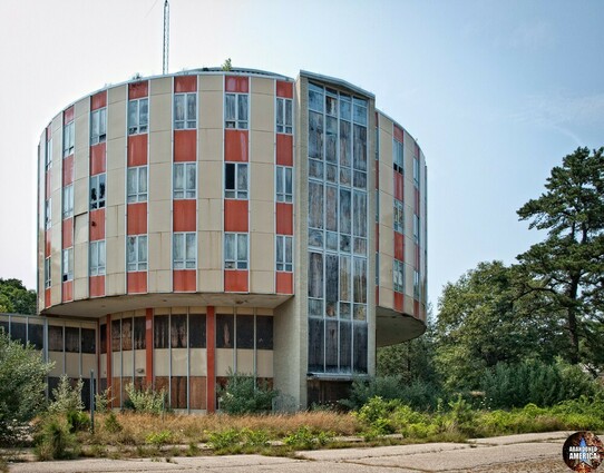 A round building with a column of windows running up one side, all boarded. Rows of windows alternating with orange panels are broken by columns of pale yellow. The plants nearby are wild and unkempt