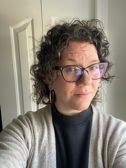 White woman with glasses, a charcoal dress and oatmeal coloured cardigan 