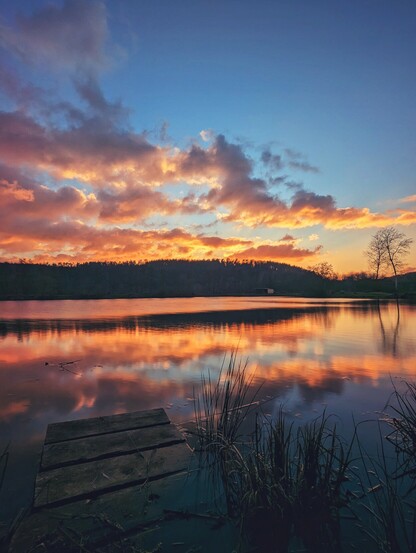 Sonnenuntergang am einem See. Reste eines untergehenden Stegs sind im Wasser zu sehen, in welchem sich der blaue Himmel mit orangenen Wolken spiegelt.