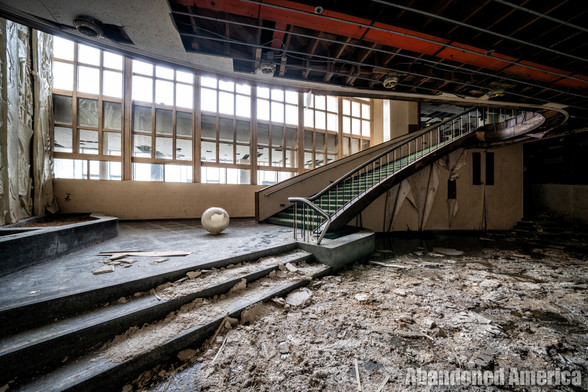 A slightly curved staircase with green steps and wood railing extends from the upper right to a landing on the lower left. A broken globe from a light rests on the landing. The floor is covered with dirt and debris. Behind the stairway a bank of long windows on the wall casts a dull light.