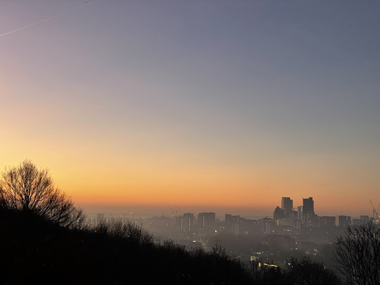 A city rises from the mist in front of a sky glowing with the sunrise colours of orange, pink and gold. 