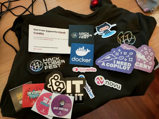 A black t-shirt with a Hacktoberfest "H"-logo. Above it are stickers scattered. Visible/Readable are novu, appwrite, devtron, and docker.