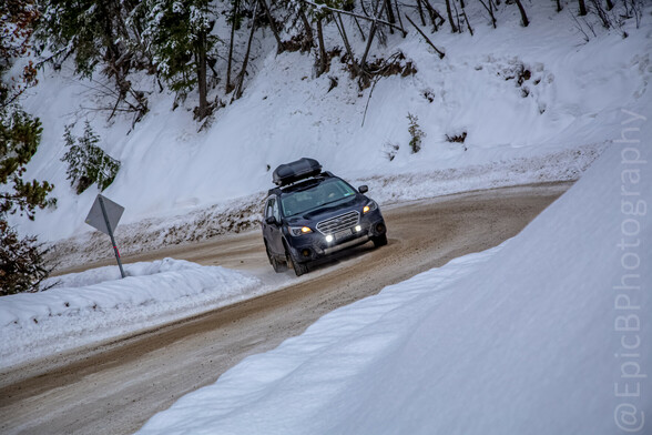 Same car, lights on, driving around a curve. The road is dirty snow, and everything else is snow covered