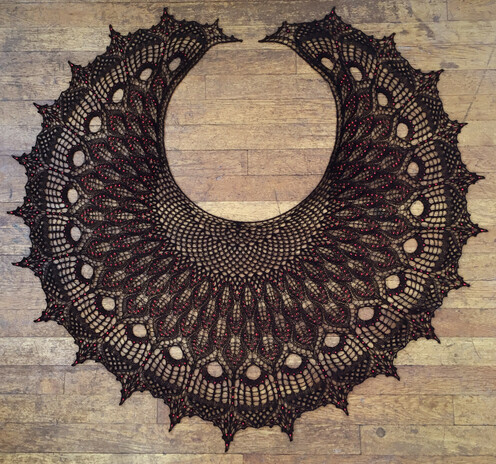 Full view of a crescent-shaped knitted lace shawl made of black yarn with many red beads, sitting on a floor of narrow wooden boards.