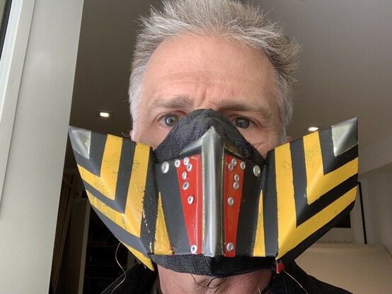 Photo of a man wearing a mask made of angular, folded and riveted sheetmetal painted red, with bold yellow and black stripes. His eyes, forehead, and grey hair protrude above the edge of the mask. 