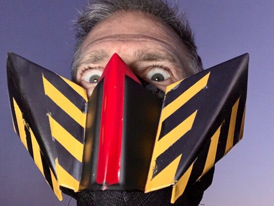 Photo of a man’s face wearing a mask made of angular, folded sheetmetal painted red, with yellow stripes. The image is shot and lit from below, looking up. The mask frames hie eyes which are wide and angry-looking in contrast with the soft dusk sky behind him. 