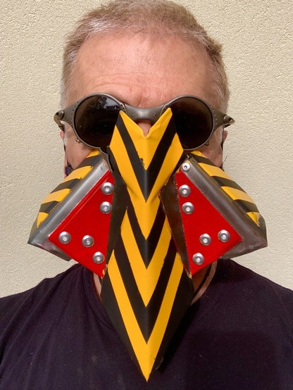 Portrait photo of a man wearing round Oakley brand sunglasses and a mask made of angular, folded and riveted sheetmetal painted with two red triangles either side of his nose and yellow and black bumblebee stripes in a chevron shape down the sharp, pointed nosepiece. The nose piece juts down from his brow line to below his chin. 