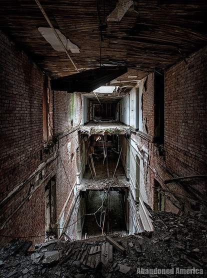 A view over a segment of hallway that has collapsed leaving the two floors below visible. The walls are brick and the ceiling is tilted and close to collapse