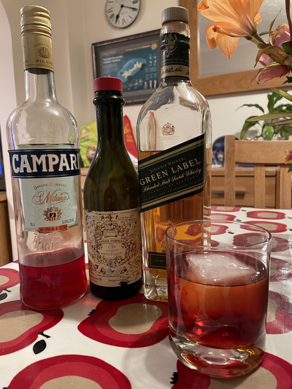 A bottle of Campari next to a bottle of Antica Formula next to a bottle of Johnnie Walker Green Label. In front is a glass with a boulevardier in it with a single large ice cube.