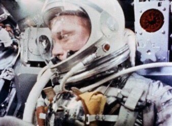 A color photo of John Glenn in the Friendship 7 capsule he used to orbit the Earth. He is wearing a spacesuit like the one worn by Shepard in the previous photo. A large, round helmet sports a translucent visor through which Glenn's brow, eyes, and nose are visible in profile. Several white tubes connect to the helmet. The suit itself is made of a silvery, reflective material. Glenn is strapped in with what appears to be a canvas harness. Numerous instruments are visible behind and around him.