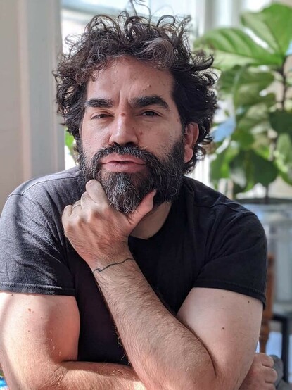 Headshot for Nathaniel Justiniano, a man with a little salt in a pepper beard and mustache, dark wavy hair, thick dark eyebrows, and a tattoo encircling his wrist. He holds his chin in the hand with the tattooed wrist and is wearing a dark t-shirt.