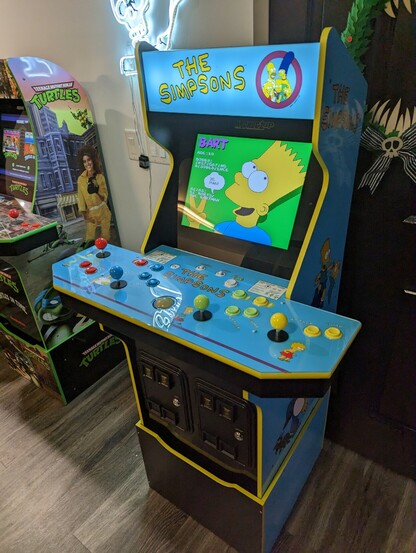 The Simpsons arcade machine is pictured. The original The Simpsons arcade game is on the screen with Bart Simpson visible.