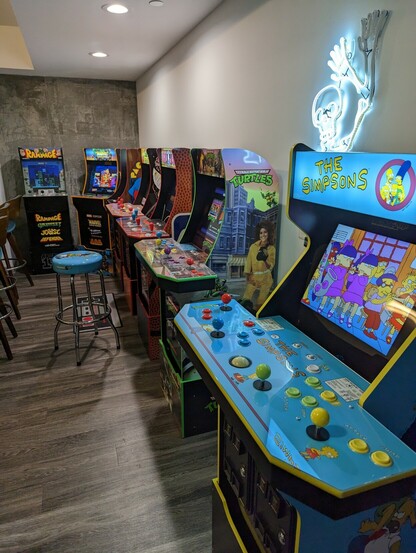 Seven arcade machines are lined up in a row. From left to right, they are Rampage, Marvel Super Heroes, Pac-Man, Golden Axe: The Revenge of Death Adder, NBA Jam, Teenage Mutant Ninja Turtles, and The Simpsons.