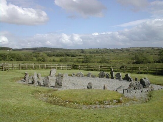 Two thirds of the view is green landscape, one third is pale blue sky. There’s a stone circle made from low, grey stones in the centre of view.

None of the stones are more than half a metre in height. The centre of the circle is covered with loose hardcore that looks very modern. Behind the circle, running horizontally in the frame of the photo is a wooden fence.

Between the spot the photo was taken from and the circle is a low, circular mound covered with green grass. An irregular stone kerb is partially visible through the longer grass growing around it.