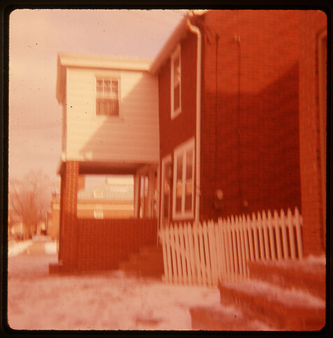 Slightly blurry and faded color transparency image looking down the street past a house with a brick porch. A white, picket fence stands with the gate open. A dusting of snow covers the brick sidewalk.