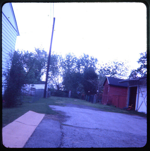 Slightly dark color transparency image, camera held at a slight angle, looking down a wide alley with garages along one side. Kids of a certain age will spot the Marx Big Wheel in the open garage to the right.