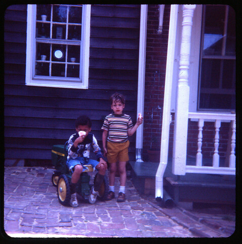 Slightly dark color transparency image of two children. One is sitting on a ridable John Deere tractor toy while the other stands. Nothing quite captures childhood in Old New Castle in this period like kids with paper cones of Rosati water ice, purchased at Wassams.