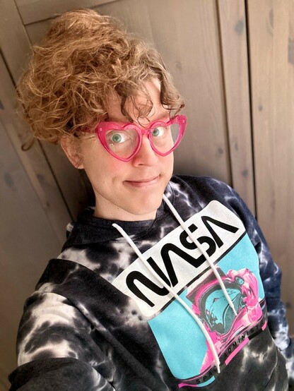 me with my hair up in a very messy pineapple, wearing my fuchsia heart-shaped glasses and a black and white NASA hoodie that has a hot pink and light blue photo of an astronaut in a space suit on the front