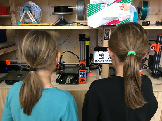 Zu sehen sind zwei Jugendliche, die vor einem 3D-Drucker stehen und den Druck beobachten.