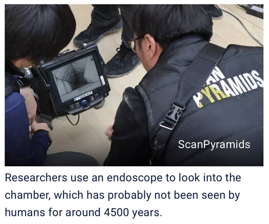 Photo of two people looking at a monitor on which the inside of a stone tunnel can be seen. One of the people is wearing a vest with "Scan Pyramids" written on the back.