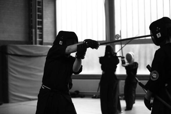 4 martial arts students in fencing gear and as armor. Two duelling in the background, two Infront. The one on the left strikes horizontally with the wooden sword to the student passively standing on the right