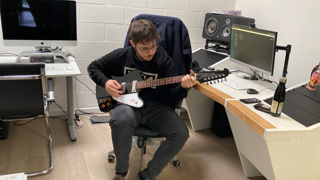 A person with glasses playing a firebird-style guitar next to his desk.