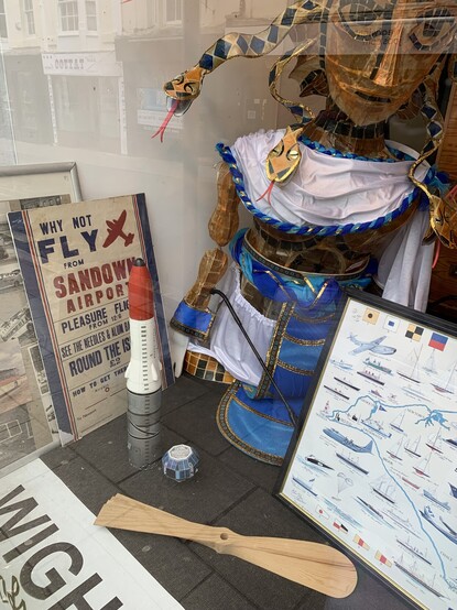 Small models of the BLACK ARROW rocket and the Prospero satellite in an ex-department store window in Ryde on the Isle of Wight. Also pictured, is a large theatre prop of Medusa, a vintage Sandown Airport poster, some old bus photographs and a framed picture of aircraft and ocean vessels.