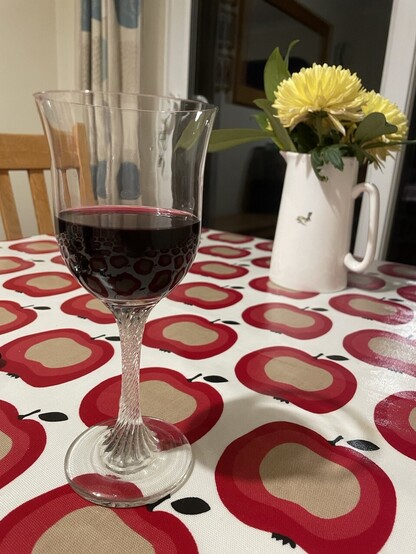 A glass of red wine sitting on a table cloth covered with a stylised red-apple print. I. The background is a tall white jug with yellow flowers in. 