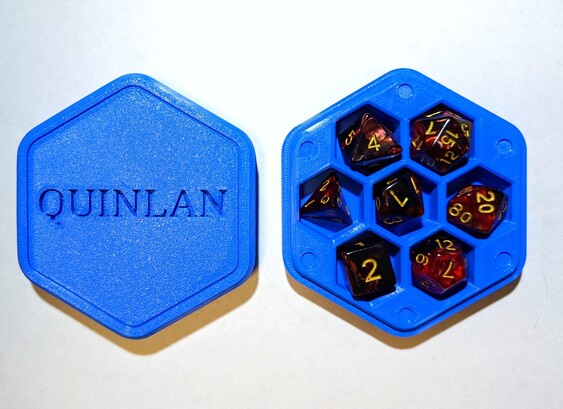 A hexagonal dice box with 7 DND-style dice in hexagonal slots. The box is blue plastic, and the name "Quinlan" is inset into the top. 