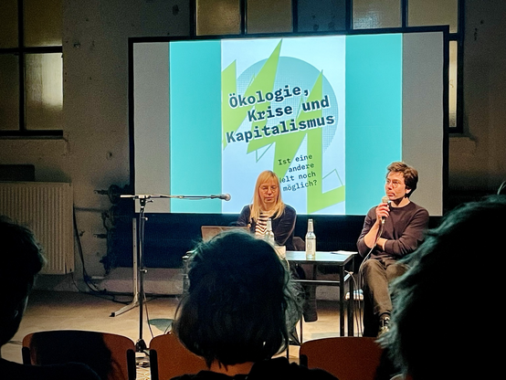 Referentin und Gastgeber sitzend vor Leinwand mit Veranstaltungsgrafik - im Vordergrund Silhouette von Teilnehmenden.