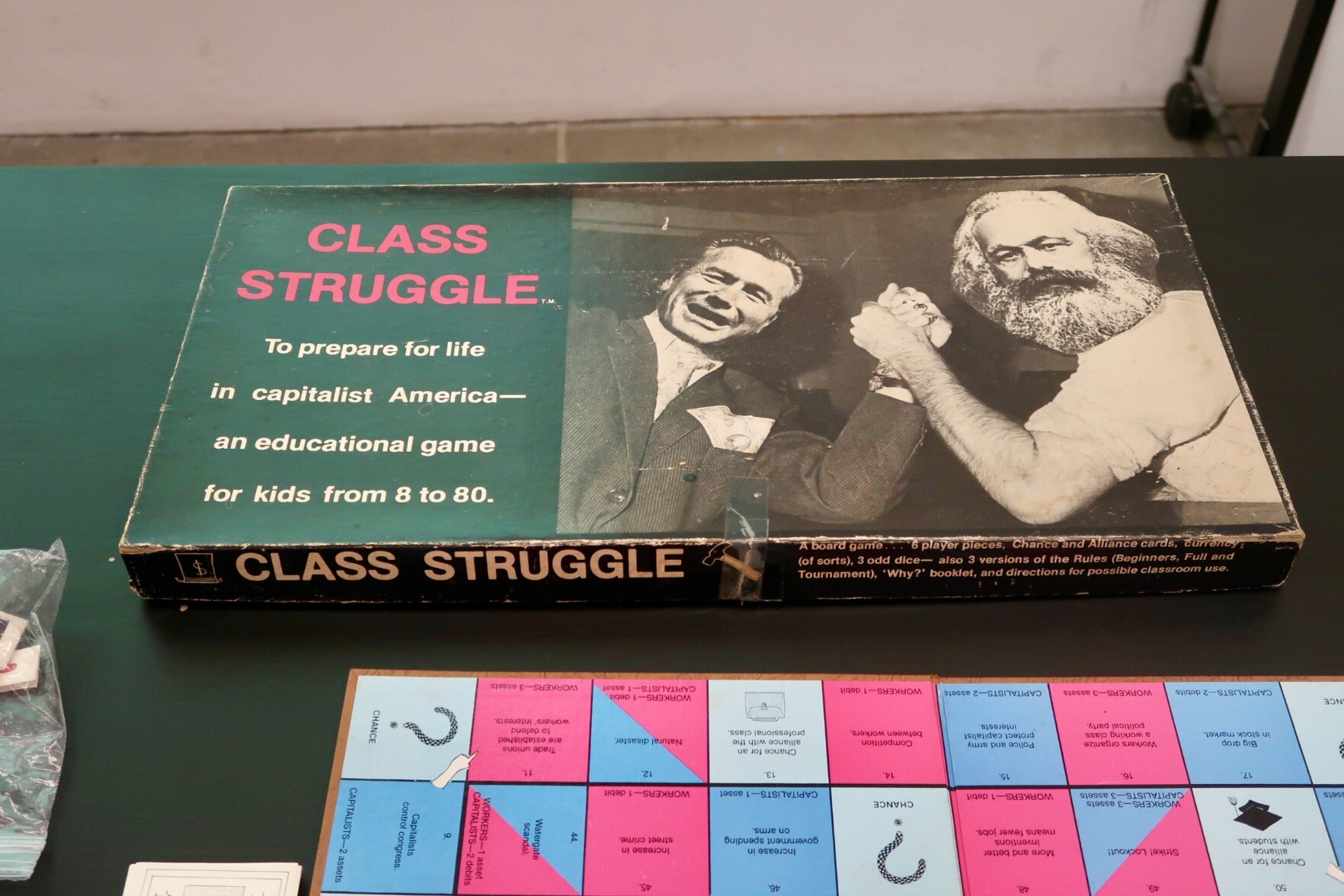 The box for Class Struggle. It depicts a black and white photo of Rockerfeller and Marx arm-wrestling. The two men are using their left arms. The text on the box says "To prepare for life in capitalist America- an educational game for kids from 8-80."