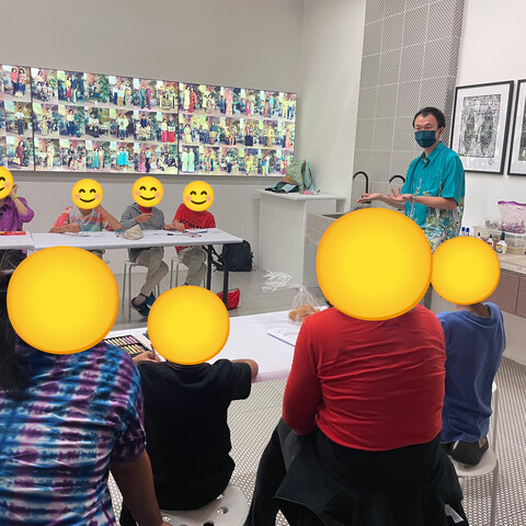 A scene from a bright indoor room with a lightbox of photos and some framed pictures on the wall. In the foreground and background are two rows of people, some adults and some children, who have their faces covered with generic smiley face emoji. In the middle is the facilitator of the group, who is standing and wearing a bright green shirt.