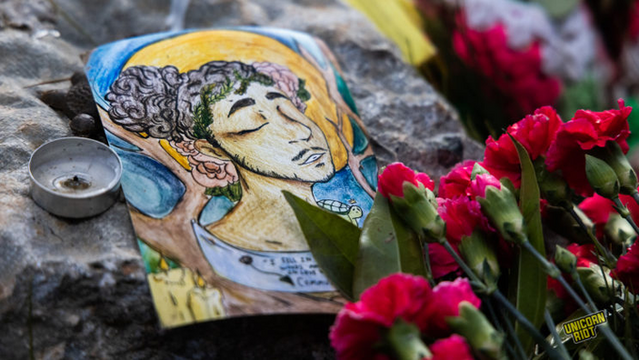 A colorful memorial drawing of murdered forest defender Manuel Teran aka 'Tortuguita' is placed upon a rock with a candle next to it and red flowers placed near the base of the rock with the drawing on it.